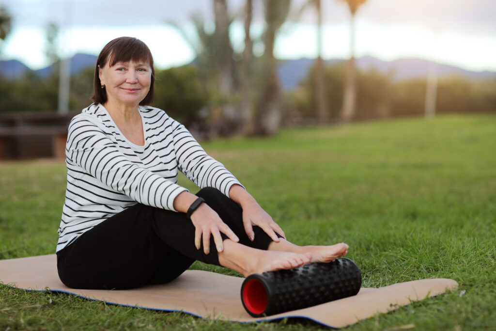 Smiling middle aged sportive woman in casual sportswear sitting on yoga mat and relaxation after workout with foam sport massage cylinder outdoors on palms trees background. World health day.